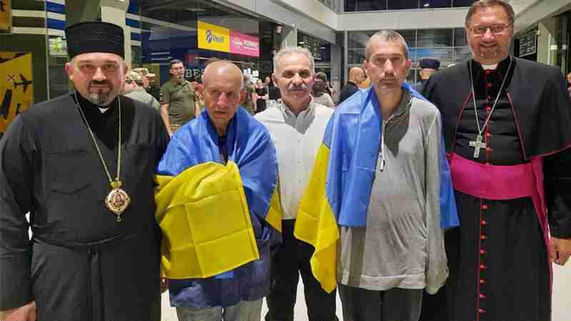 Draped in Ukrainian flags, Redemptorist Fathers Bohdan Geleta, left, and Ivan Levytsky, right, pose for a photo at the Kyiv airport in Ukraine June 29, 2024. To the far right is Archbishop Visvaldas Kulbokas, apostolic nuncio to Ukraine., tags: priester und - CNS photo/Courtesy Ukrainian Greek Catholic Church Communications Department